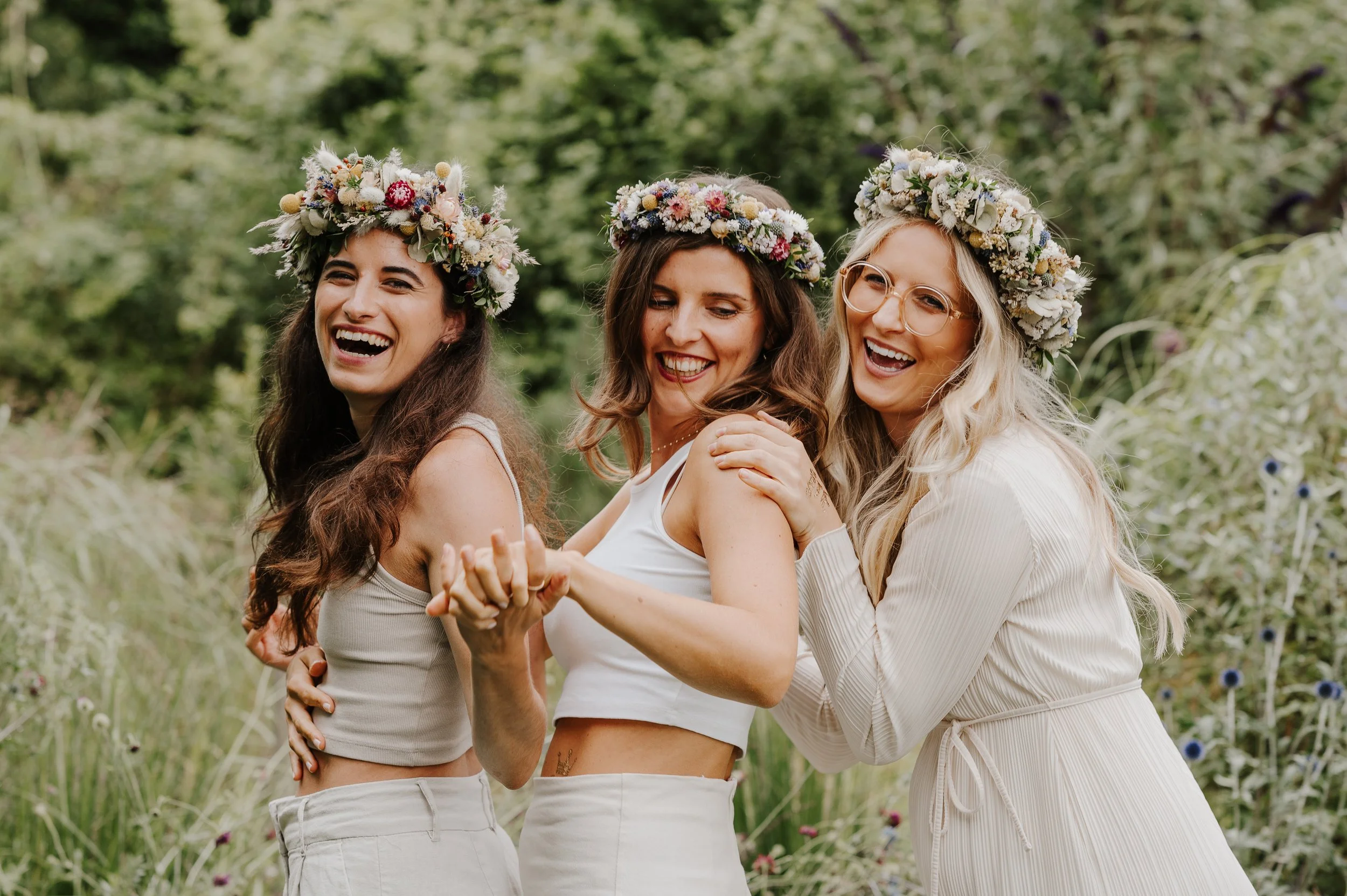 Drei lachende Frauen mit Blumenkränzen auf dem Kopf stehen im Grünen, halten sich an den Händen und umarmen sich.