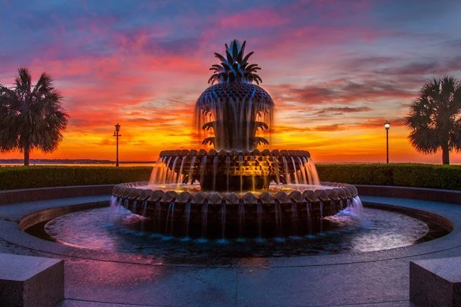 Pineapple-shaped fountain with water cascading, set against a colorful sunset sky with palm trees and lampposts nearby.