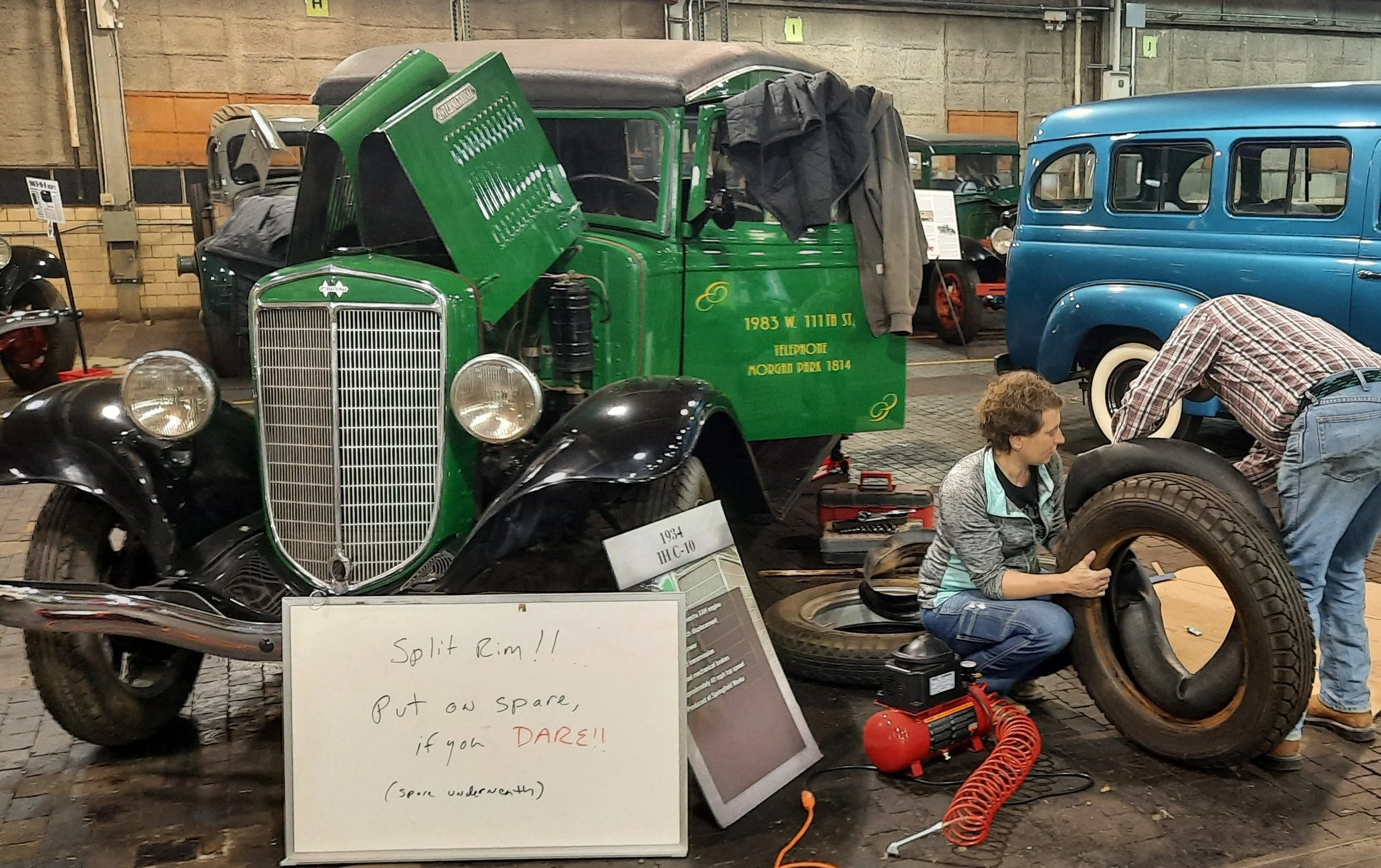 With a background of IH trucks, Sarah is sitting on the ground changing a split rim tire. There are various tools and signs around, including a whiteboard with handwritten notes about the vehicles.
