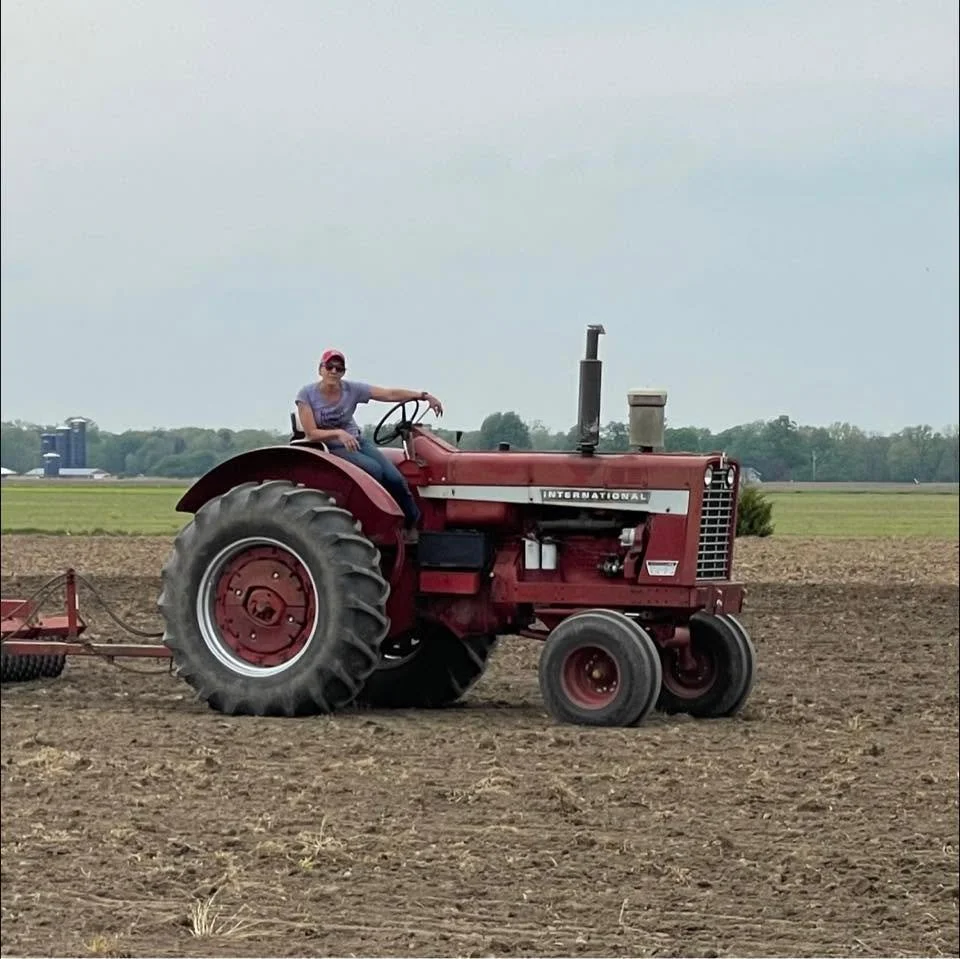 Sarah sitting on a red International tractor in a field with tilled soil, with a farm landscape in the background.