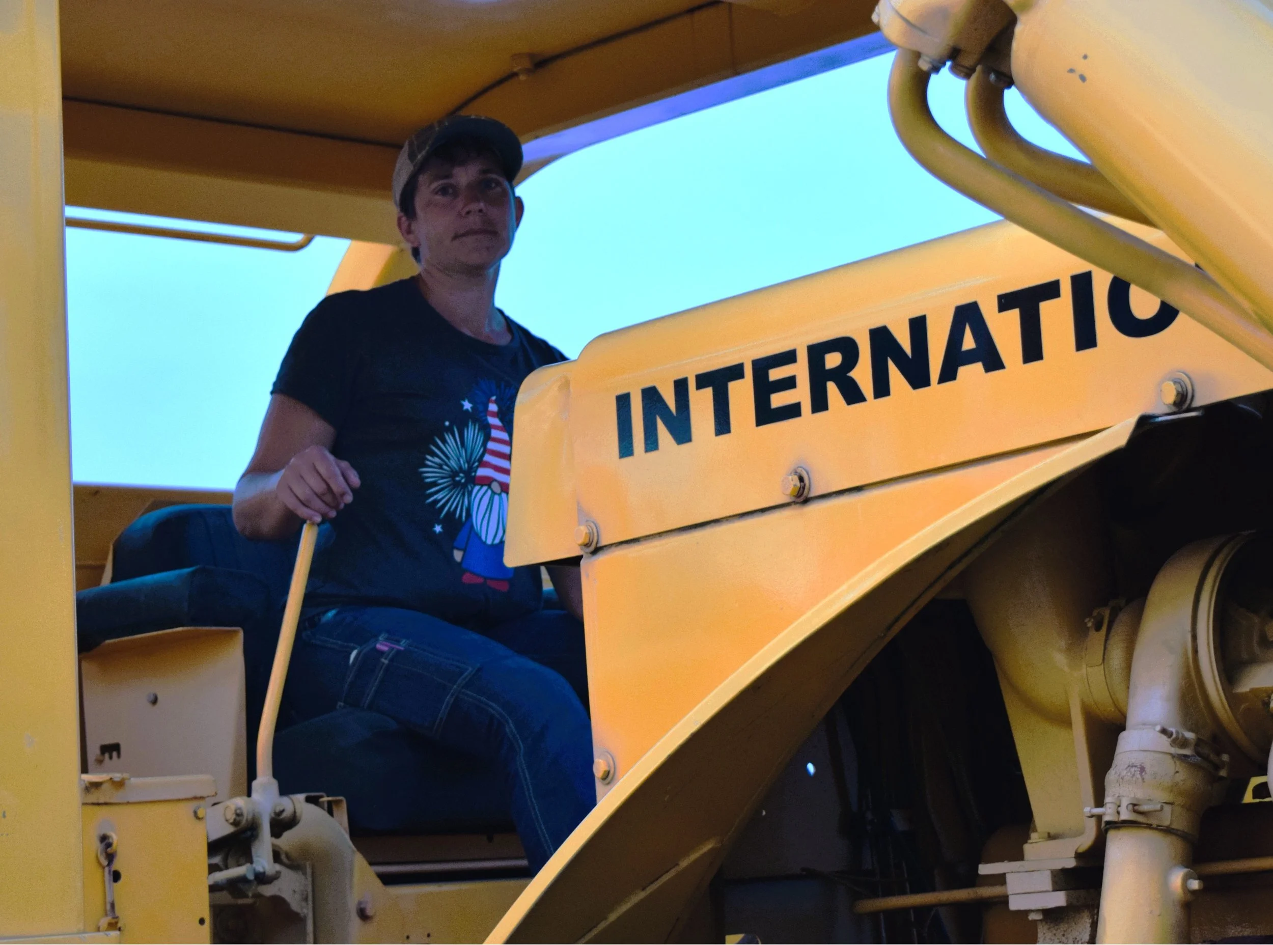 Sarah sitting in a yellow construction vehicle, holding onto a yellow handrail, with part of a sign visible that reads 'INTERNATIONAL'.