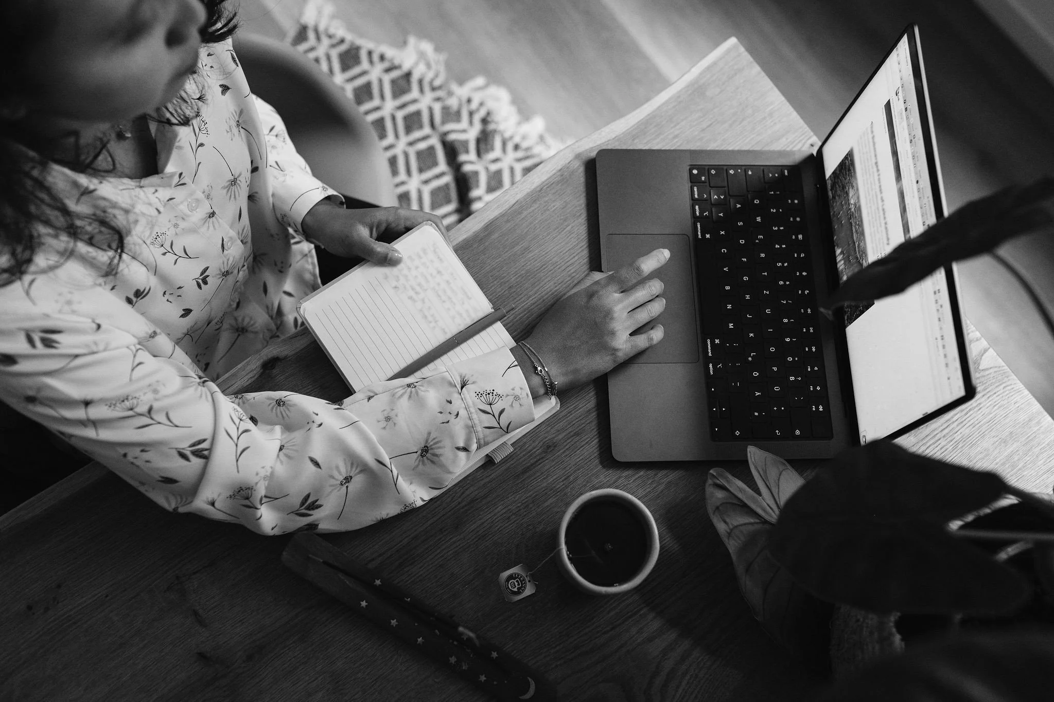Top-down view of a woman in a floral shirt sitting at a wooden table with a laptop, a cup of coffee, a notebook, and a pen.