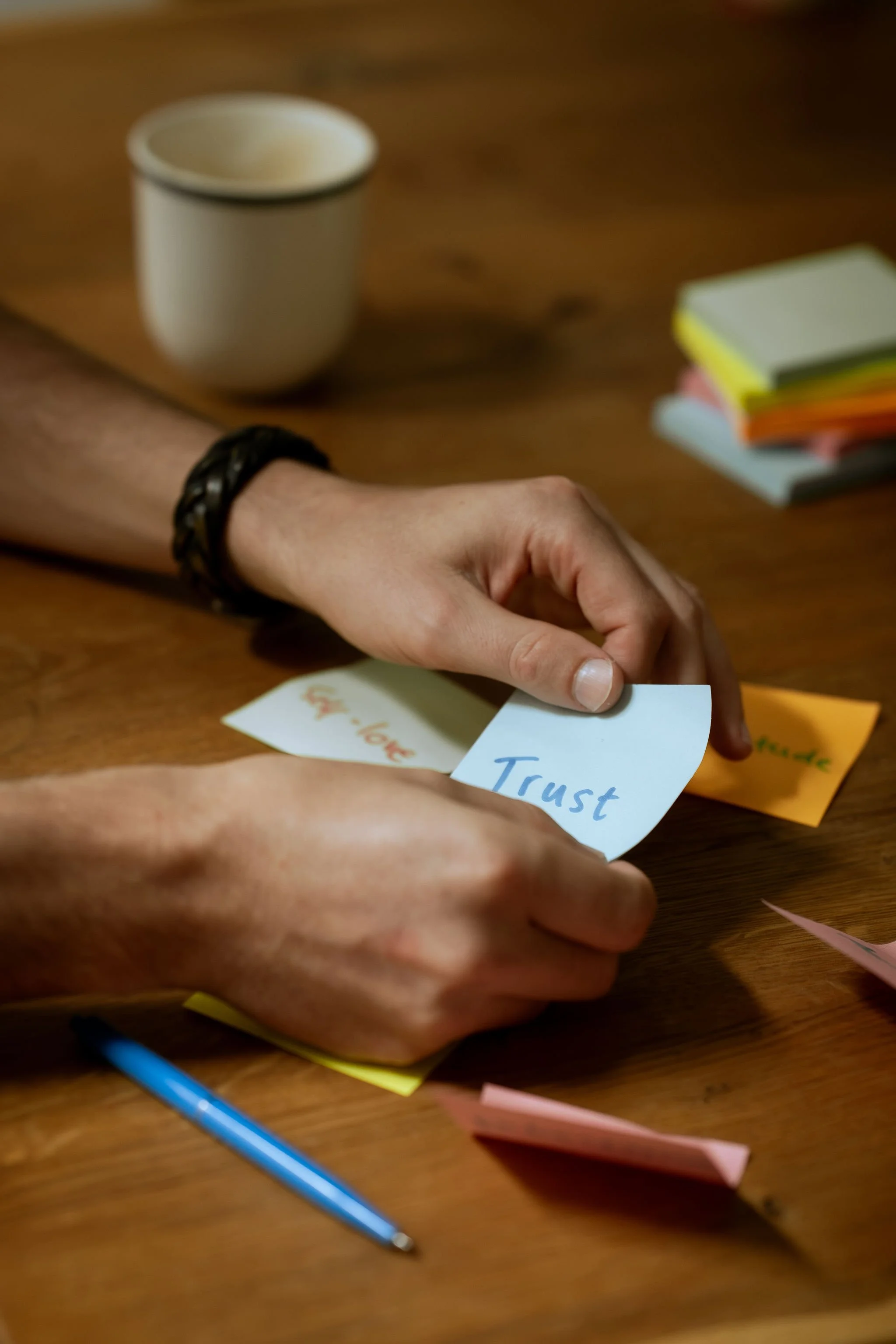 Close-up of a hand holding a blue sticky note labeled “Trust.” Other sticky notes, a cup, and a blue pen are scattered across the table.