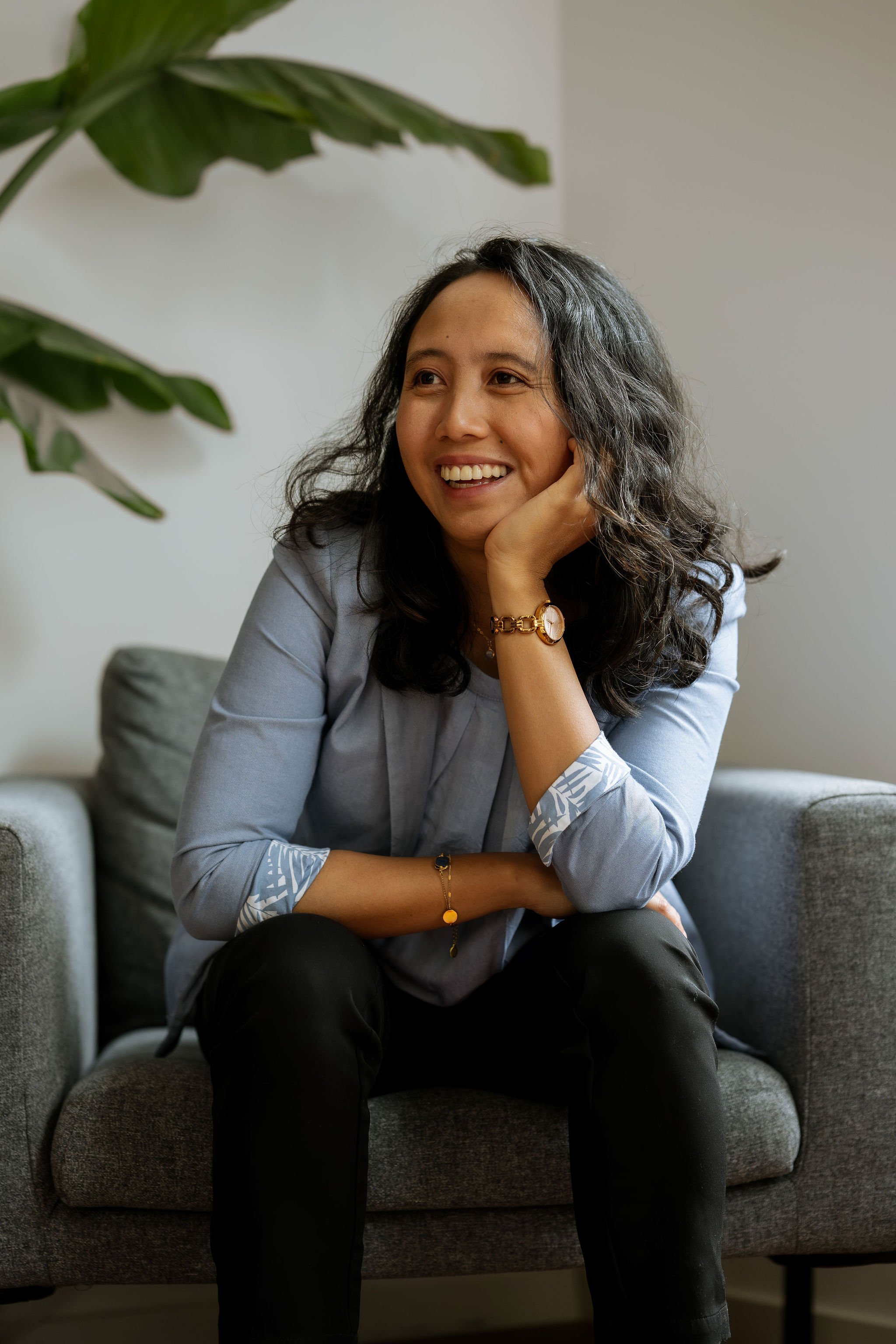 Raisa, life coach, smiling and sitting on a gray couch in a light blue blazer and black pants, resting her head on her hand in a warm, relaxed pose.