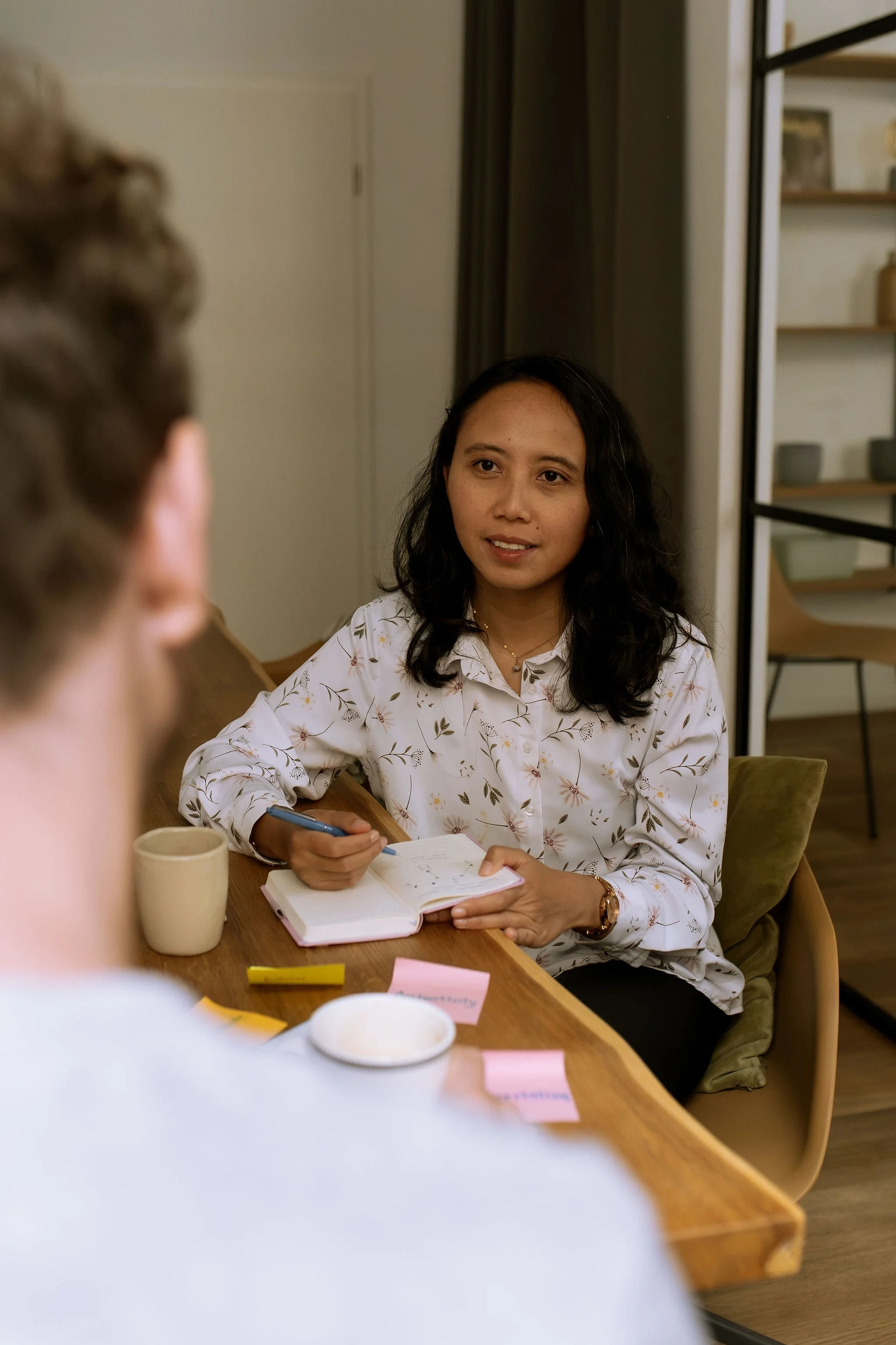 Life coach Raisa taking notes in a notebook while speaking with a client at a wooden table, with sticky notes and a mug on the table.