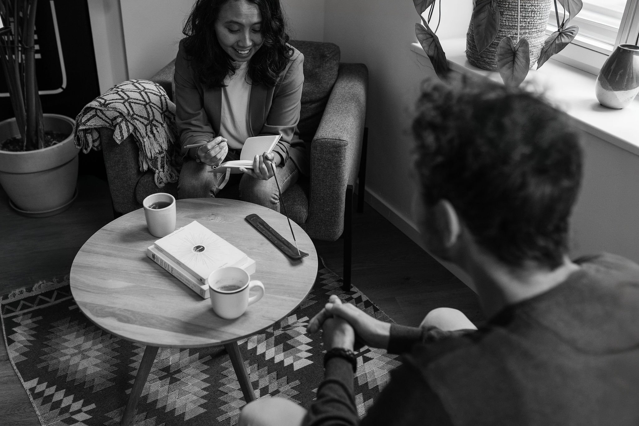 A life coach holding a pen and notebook during a coaching session with a client seated opposite her, with a table, book, and two cups between them.