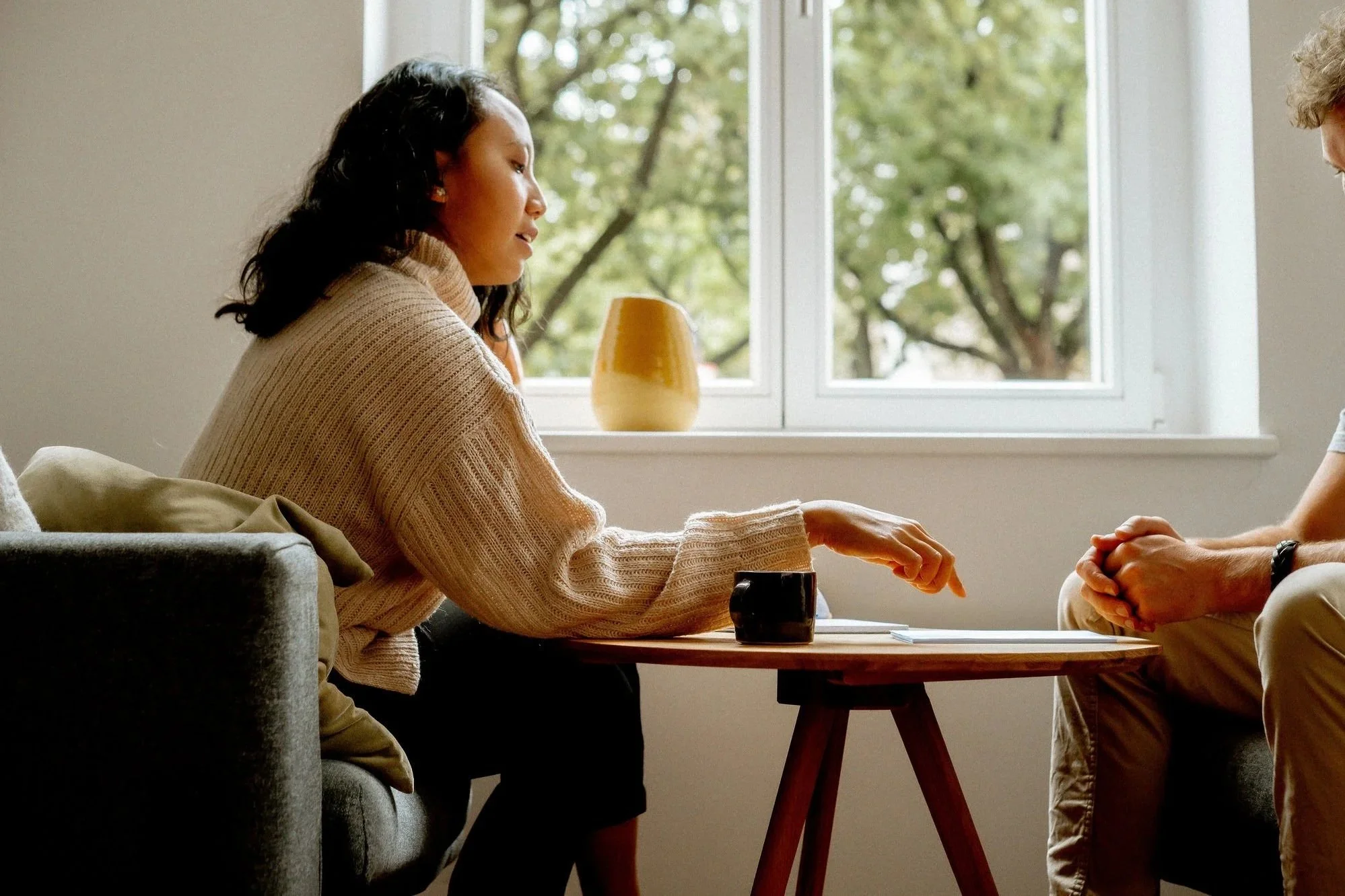 Life coach Raisa having a life coaching session with a client across a small wooden table, with greenery visible through a large window.