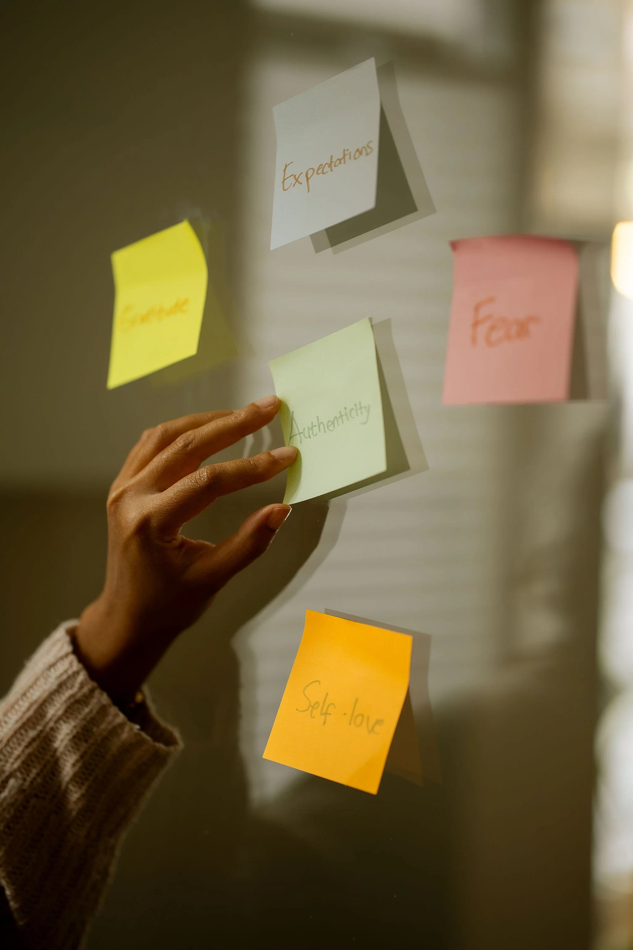 A hand placing a green sticky note labeled “Authenticity” on a wall, surrounded by sticky notes labeled “Self-love,” “Expectations,” and “Fear.”