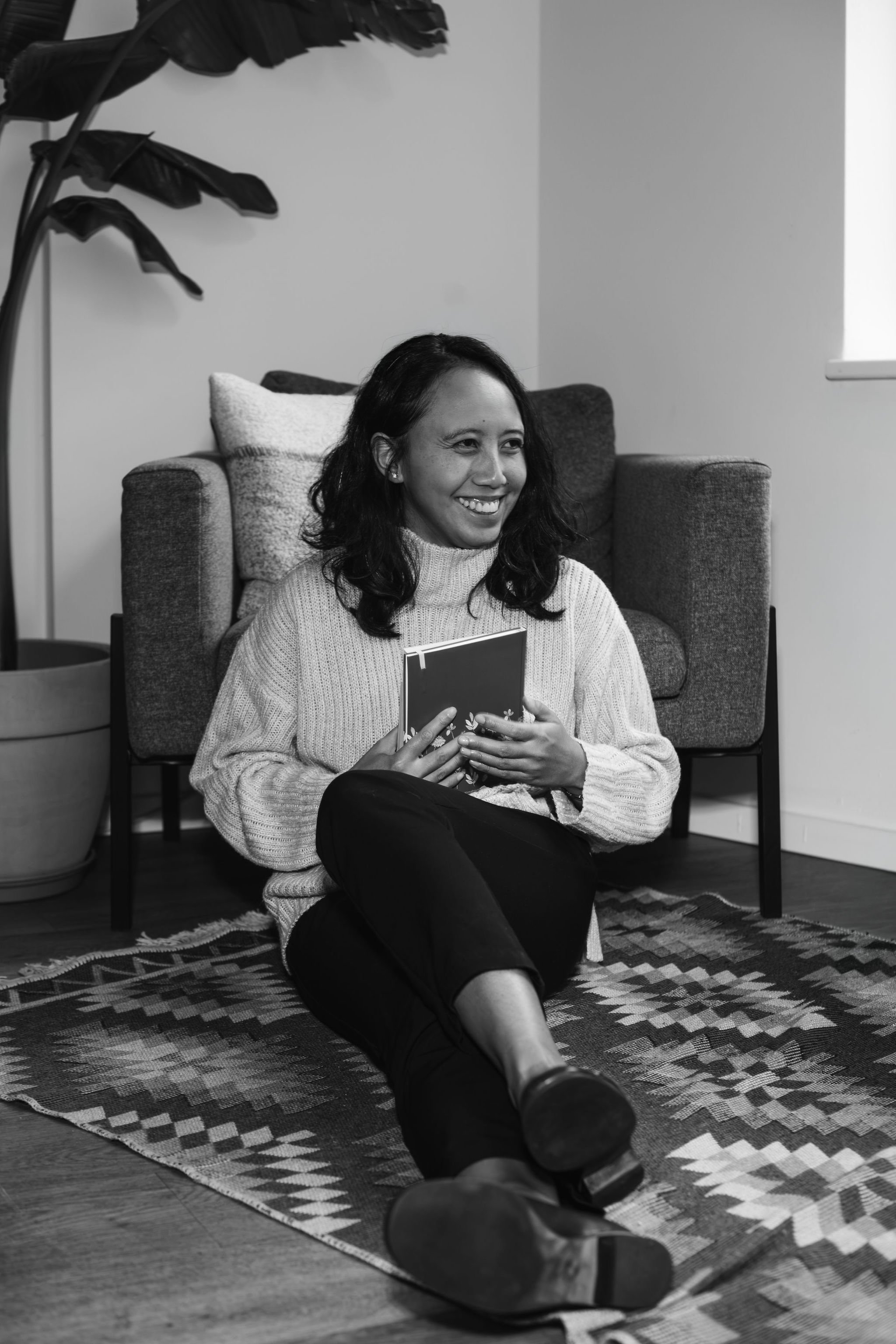 Black-and-white photo of Raisa, life coach, sitting cross-legged on a patterned rug, smiling and holding a book in a naturally lit living room.