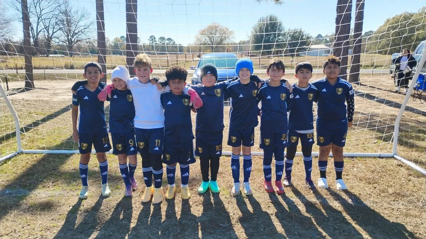 Young boys in soccer uniforms standing in front of a soccer goal on a field, posing for a team photo on a sunny day.