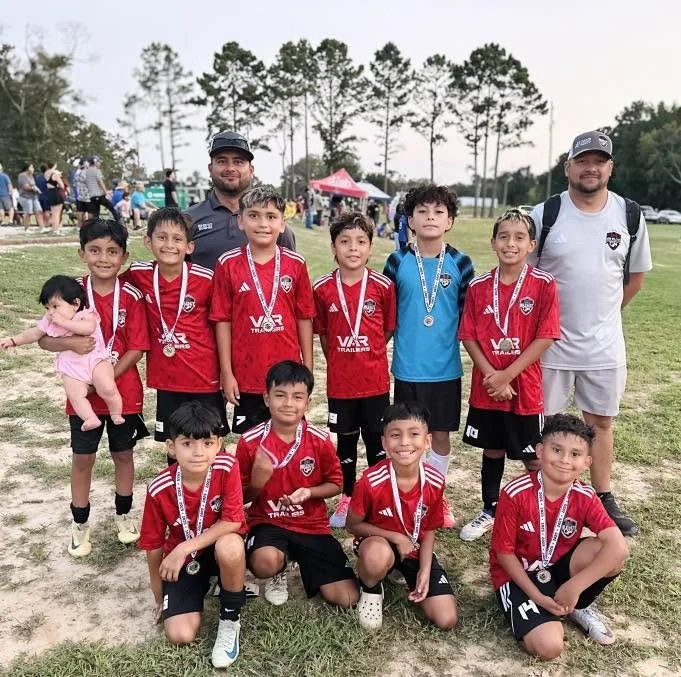 Youth soccer team posing with medals, wearing red jerseys, two coaches standing behind, fields and trees in background.