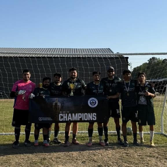 Group of soccer players celebrating with a trophy and a banner that reads 'Champions' after winning a match, standing in front of a soccer goal on a grassy field.