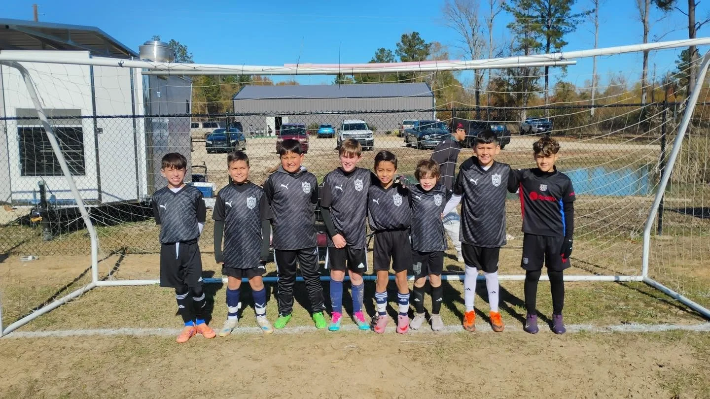 Group of young boys in black soccer uniforms standing in front of a soccer goal on a dirt field, with vehicles and a building in the background on a sunny day.
