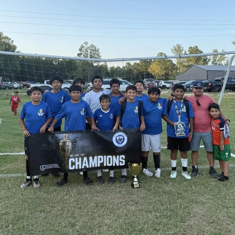 Group of young boys in blue soccer jerseys holding a banner and posing with trophies on a soccer field after winning a championship.