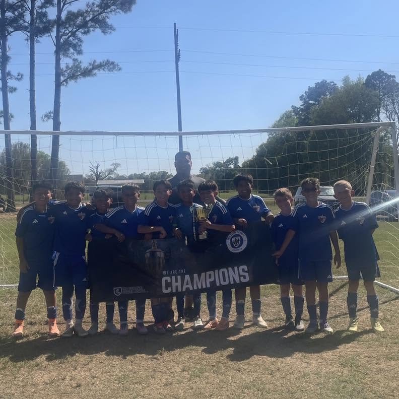Youth soccer team wearing blue jerseys holding a banner that says 'We Are The Champions' in front of a soccer goal on a grassy field.