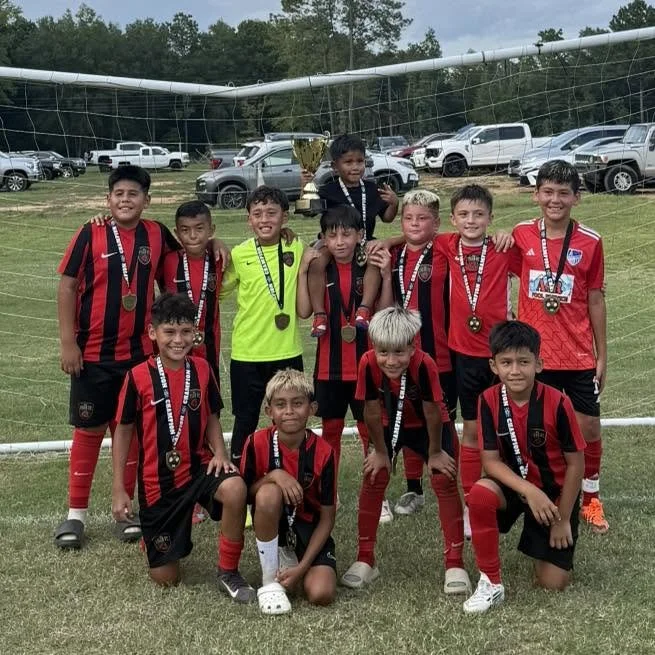 Group of young boys wearing red and black soccer uniforms posing for a team photo with medals after a match, standing in front of a soccer goal on a grassy field, with cars parked in the background.