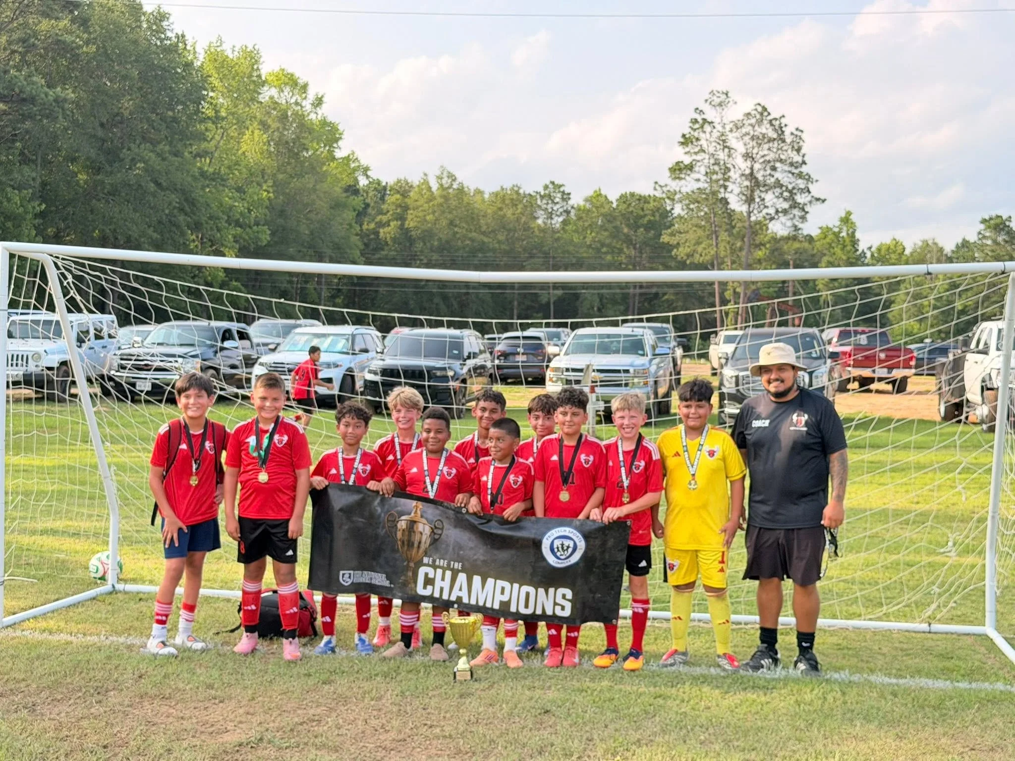 A group of young soccer players and a coach standing on a soccer field behind a goal net, holding a black banner that says 'We Are the Champions,' with the trophy placed at the center of the group, and a parking lot with cars in the background.
