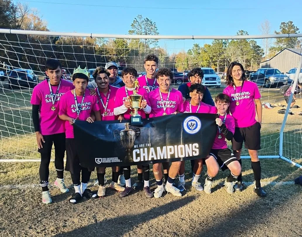 Youth soccer team wearing pink shirts holding a trophy and medals, standing behind a banner that says 'WE ARE THE CHAMPIONS', on a soccer field with a goalpost and trees in the background.