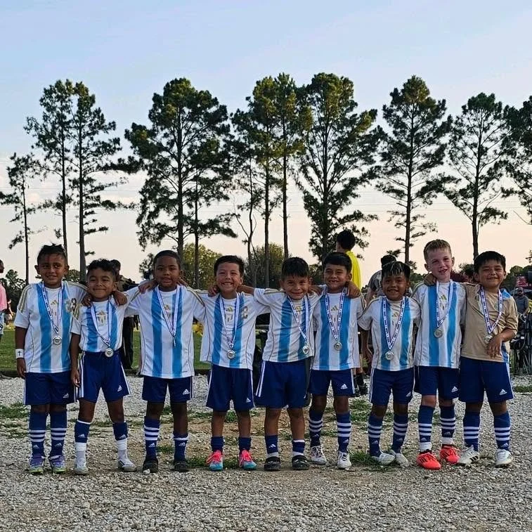 Children in soccer uniforms with medals around their necks, standing outdoors on a gravel field with trees in the background.