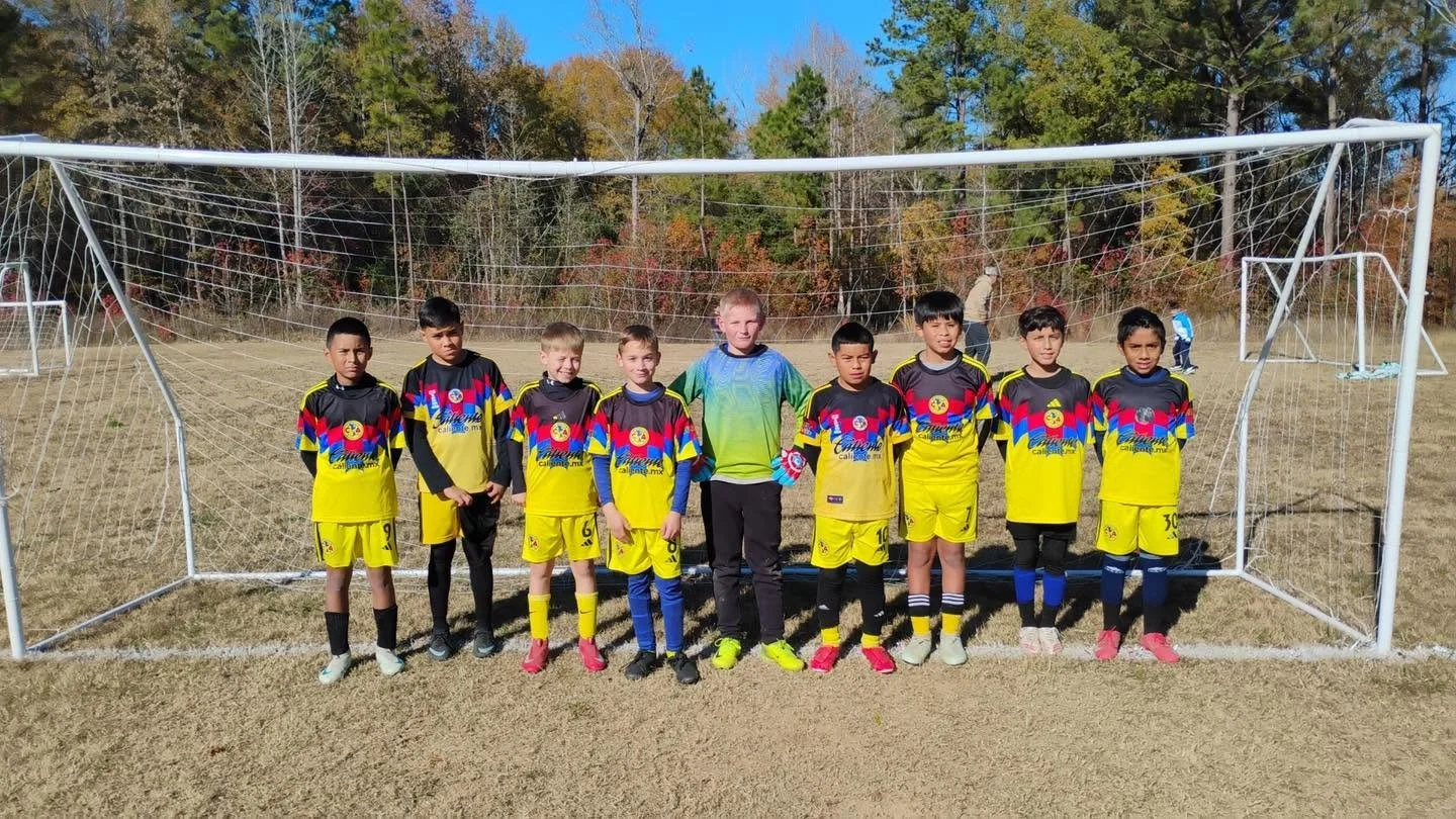 Group of young boys in soccer uniforms standing in front of a soccer goal on a field with fall trees in the background.