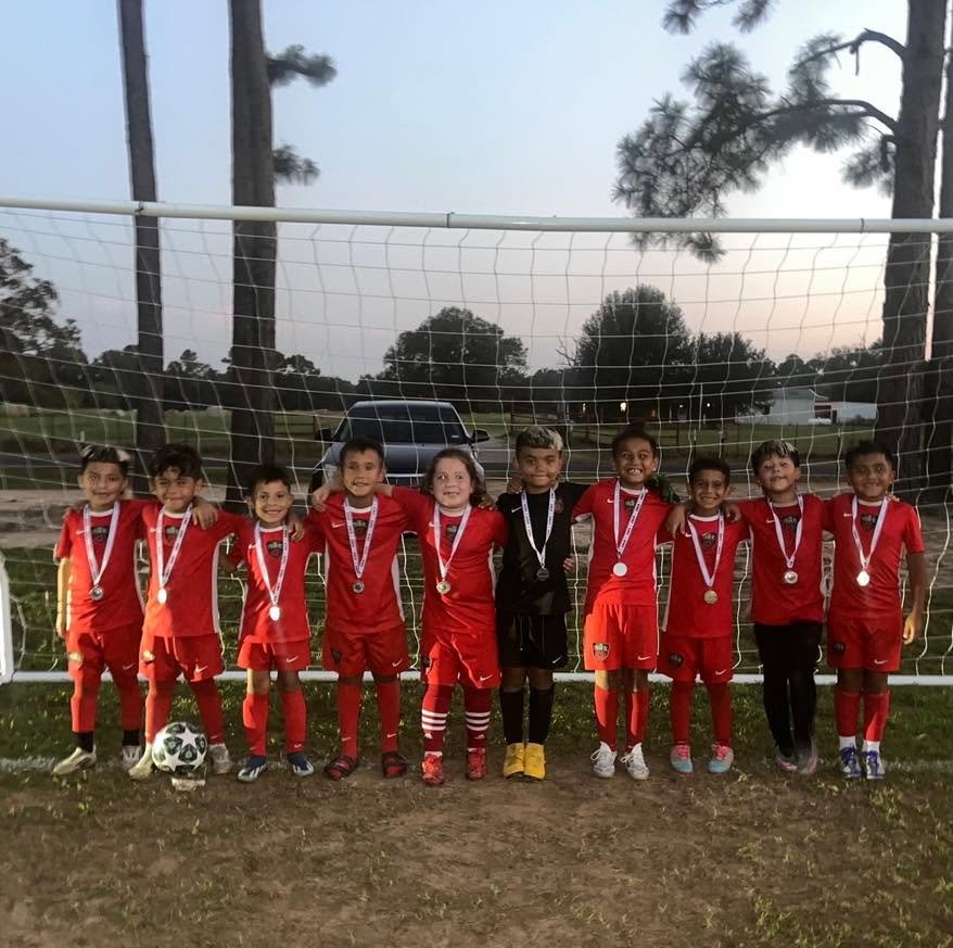 Young kids in red soccer uniforms standing in line with medals on their necks on a soccer field, posing for a team photo behind a goal post during dusk.