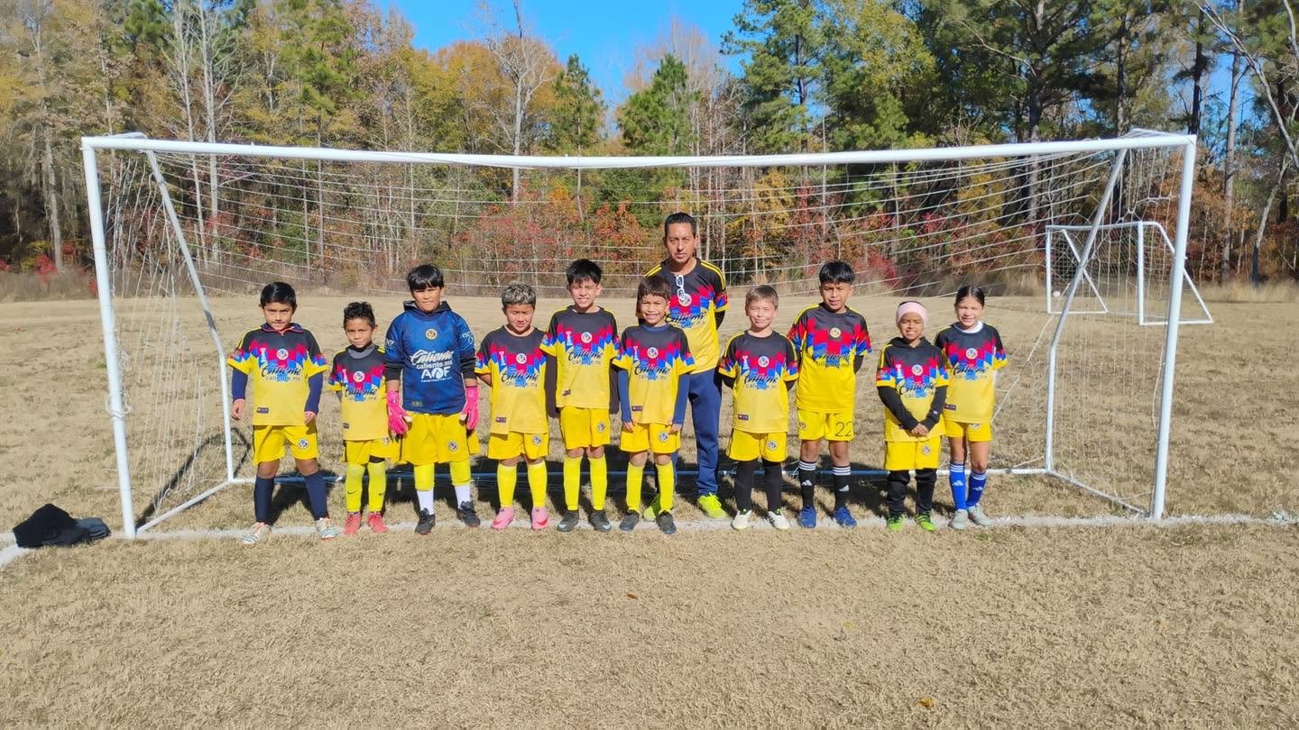 A youth soccer team standing in front of a goal on a grassy field with trees in the background. The team includes young boys and girls in colorful uniforms set against a sunny, fall day.