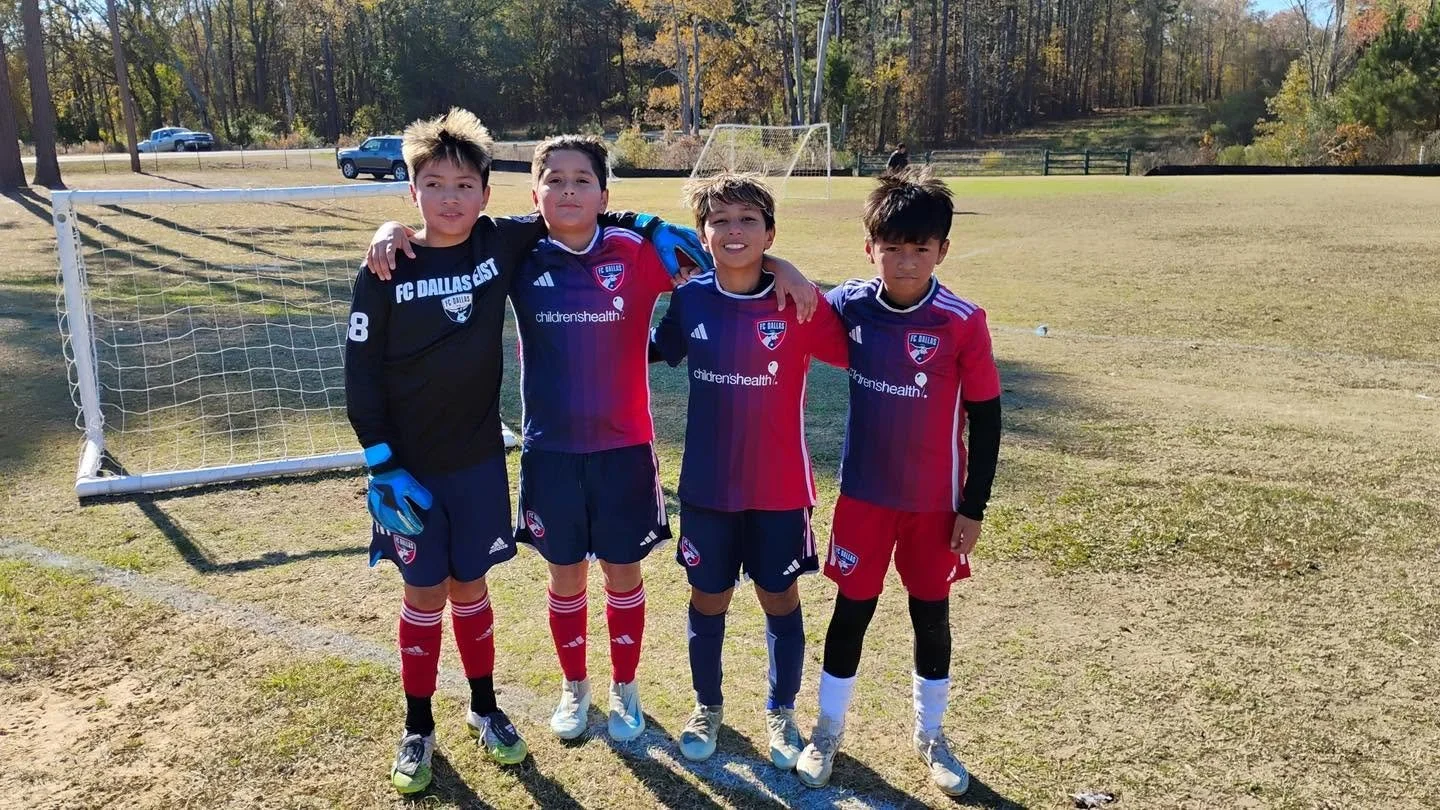 Four young boys in soccer uniforms, standing arm-in-arm on a grass field with a soccer goal behind them, posing for a photo during daytime.