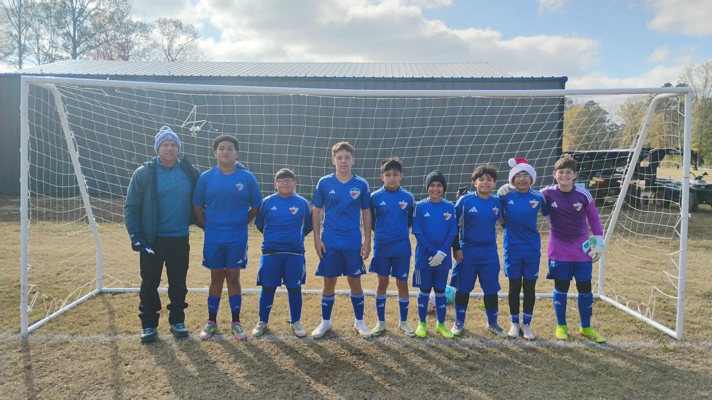 A youth soccer team in blue uniforms standing in front of a goal on a grassy field, with a coach on the left. Some players are wearing winter hats, indicating cold weather.