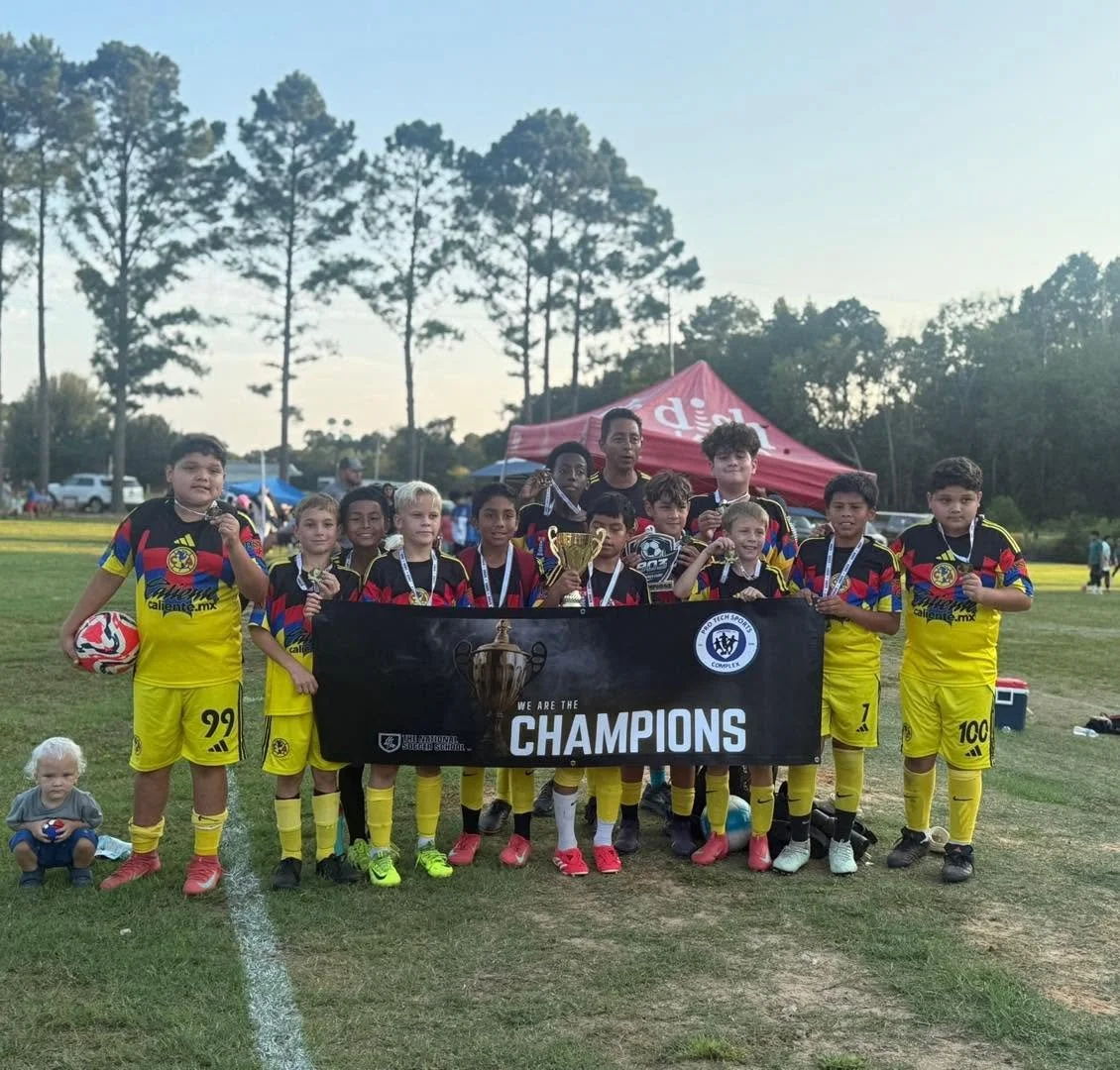 Youth soccer team in yellow uniforms holding a banner that reads 'We are the Champions' on a soccer field, with a trophy, medals, and a soccer ball, celebrating a victory.
