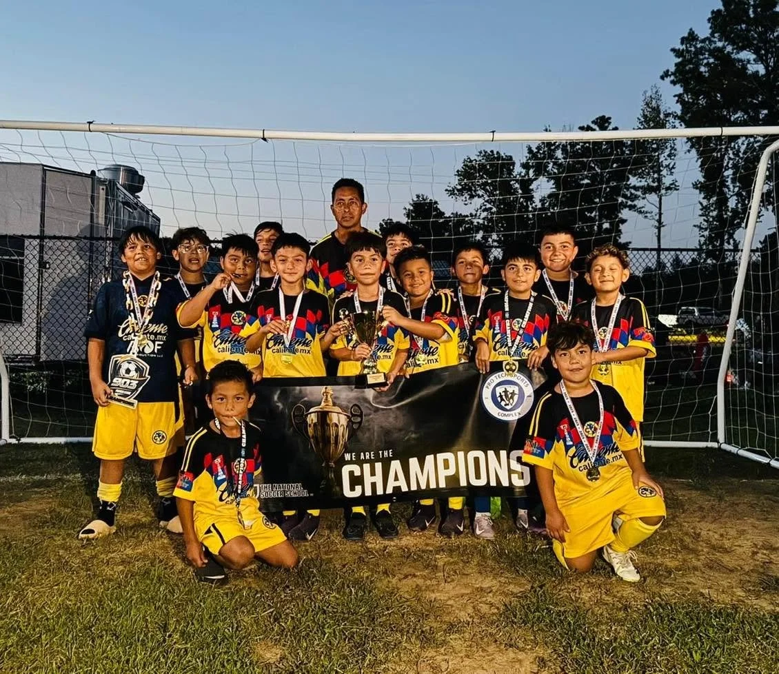 A group of young boys in yellow and black soccer uniforms standing on a soccer field, holding medals and trophies, celebrating their championship win.