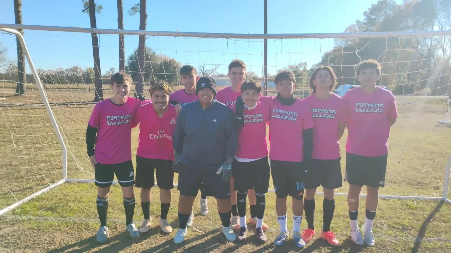 A group of nine young soccer players standing together on a field, wearing pink jerseys, with a soccer goal in the background.