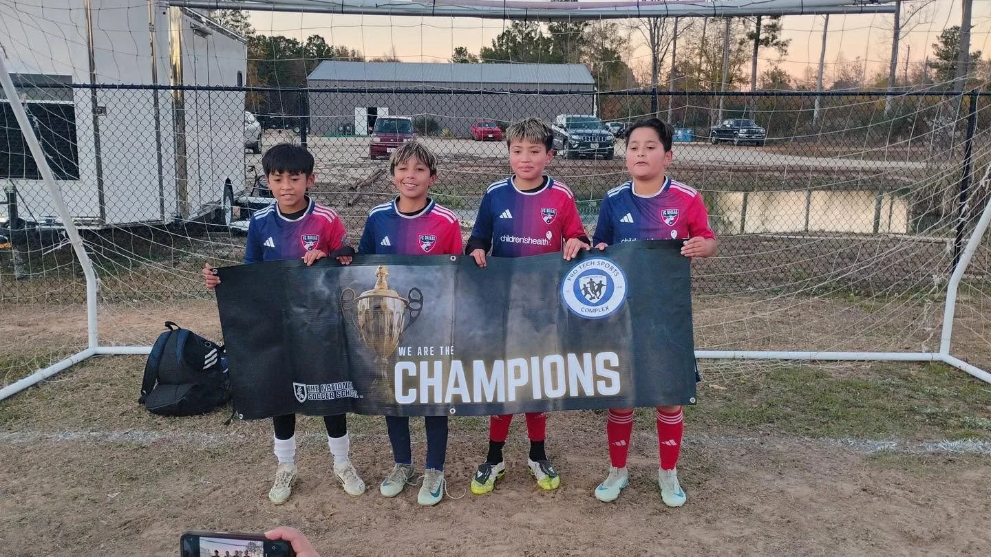 Four young boys in soccer uniforms standing in front of a goal, holding a banner that reads 'We are the Champions' and features a trophy and team logos.