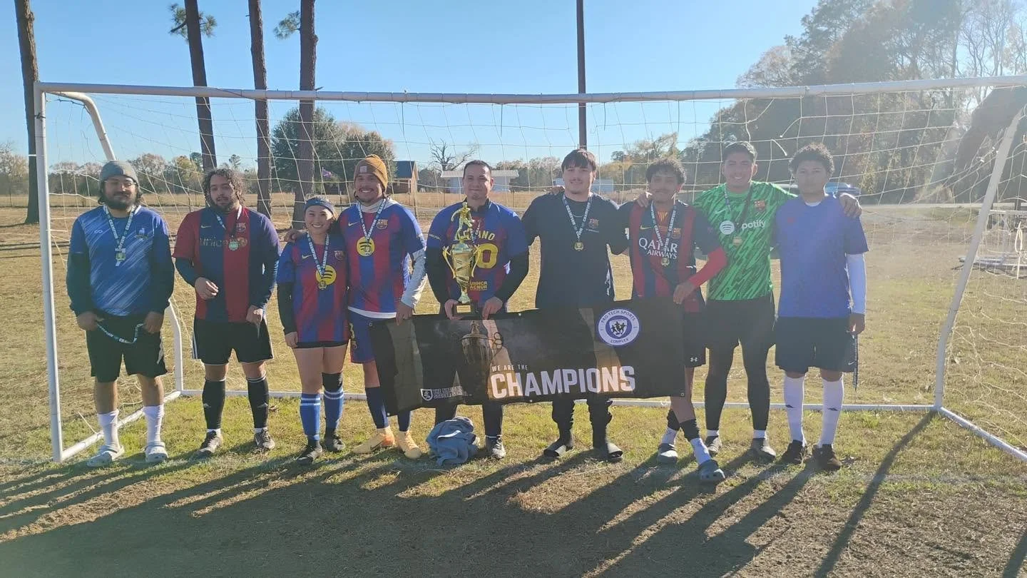 Group of young soccer players and coaches celebrating after winning a championship, holding a trophy and a 'We Are the Champions' banner in front of a soccer goal on a sunny field.