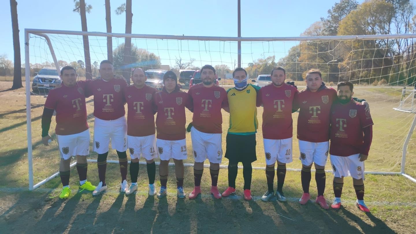 A group of nine male soccer players standing in front of a soccer goal on a field, all wearing maroon jerseys with white shorts, and one player in a yellow goalkeeper jersey. They are outdoors on a sunny day, with cars parked in the background and tr