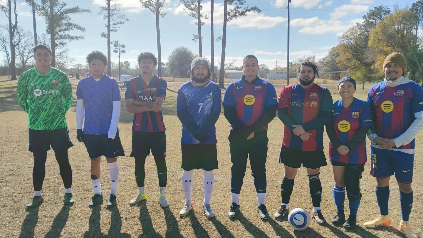 Group of nine diverse soccer players standing on a field, some wearing FC Barcelona jerseys, with a soccer ball on the ground in front of them, outdoors under a partly cloudy sky.