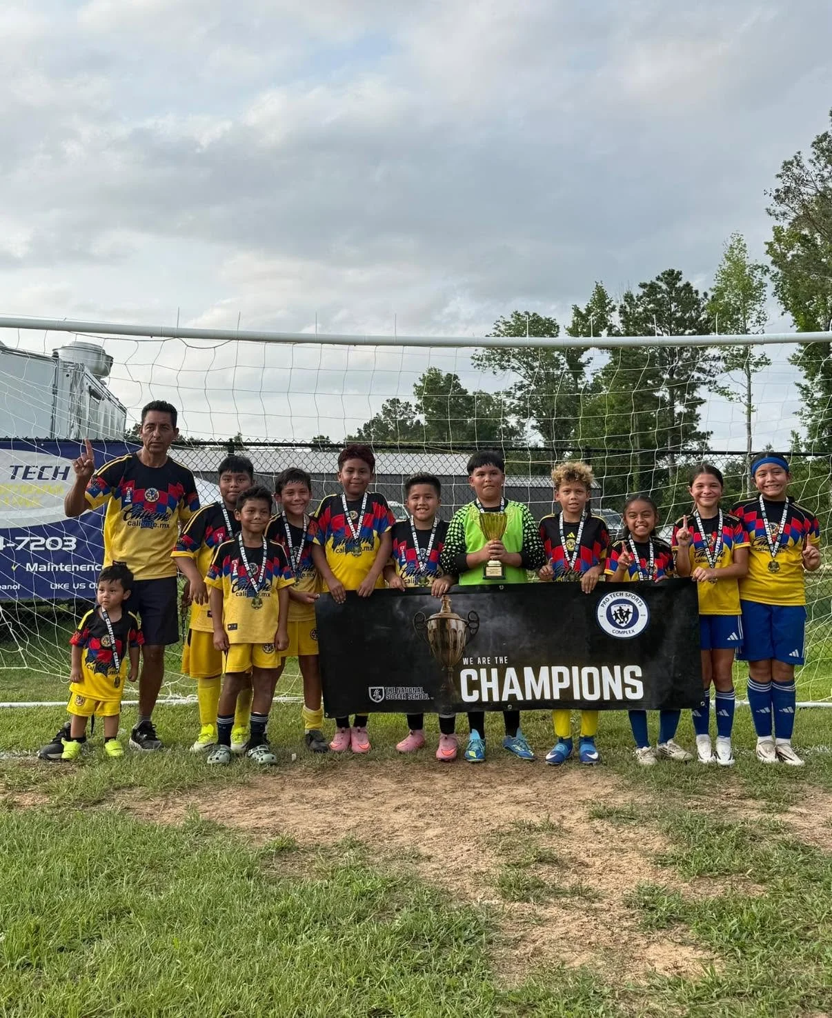 A group of young boys and girls in soccer uniforms with their coach, holding a large trophy and a banner that says 'We are the Champions,' standing in front of a soccer goal on a grassy field.