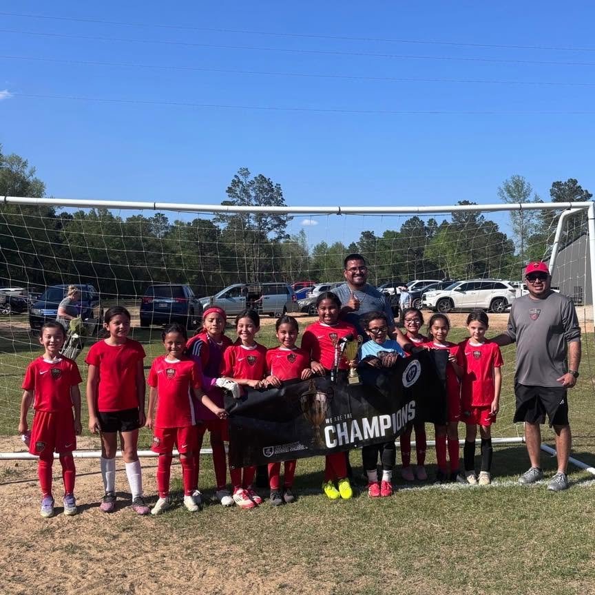 A girls' soccer team in red uniforms, along with two coaches, holding a banner that says "We Are The Champions," standing in front of a soccer goal on a grassy field, with cars and trees in the background.