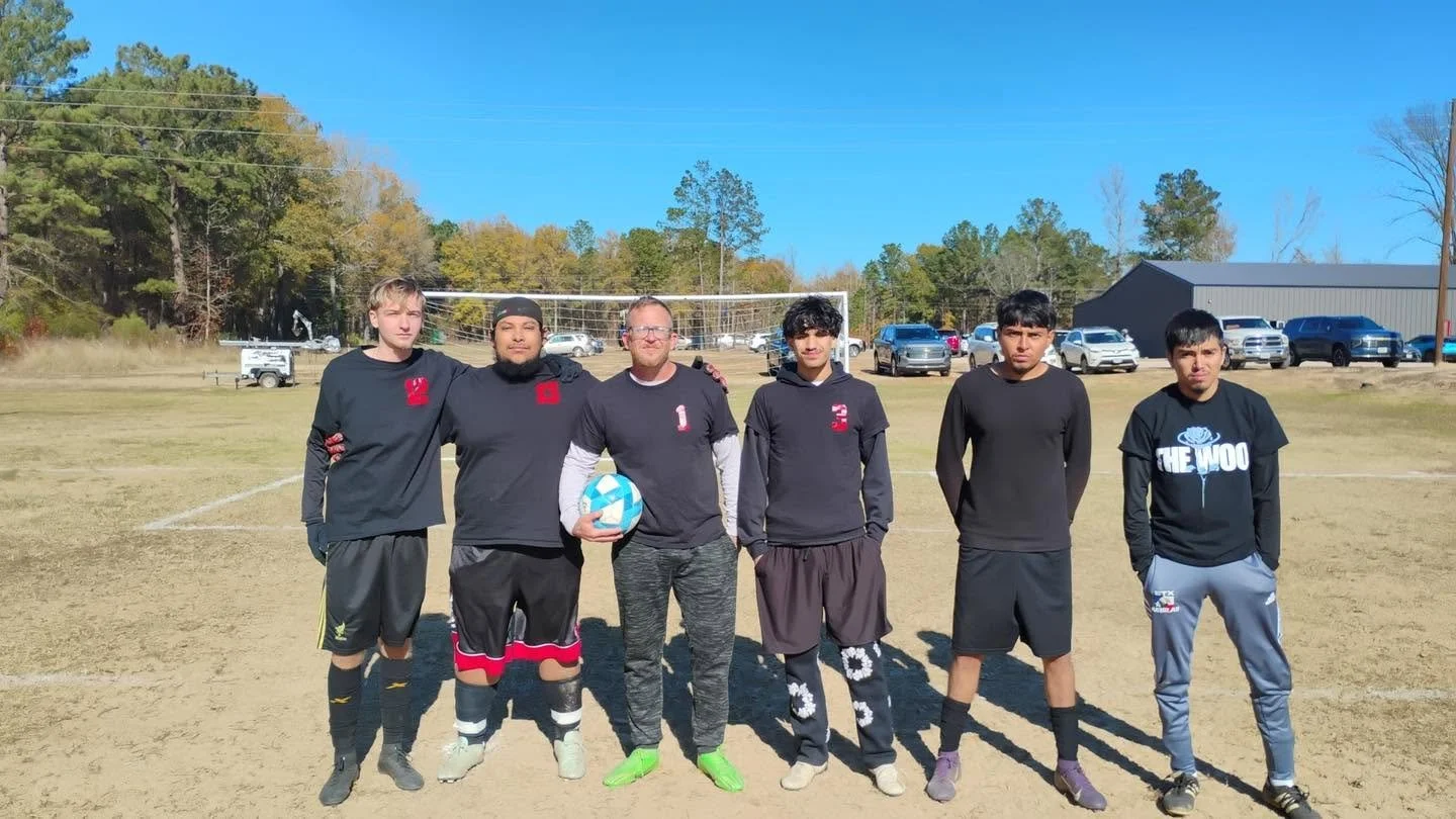 Group of six young men standing on a dirt soccer field, some wearing athletic clothes, holding a blue and white volleyball, with a soccer goal behind them and parked cars in the background.