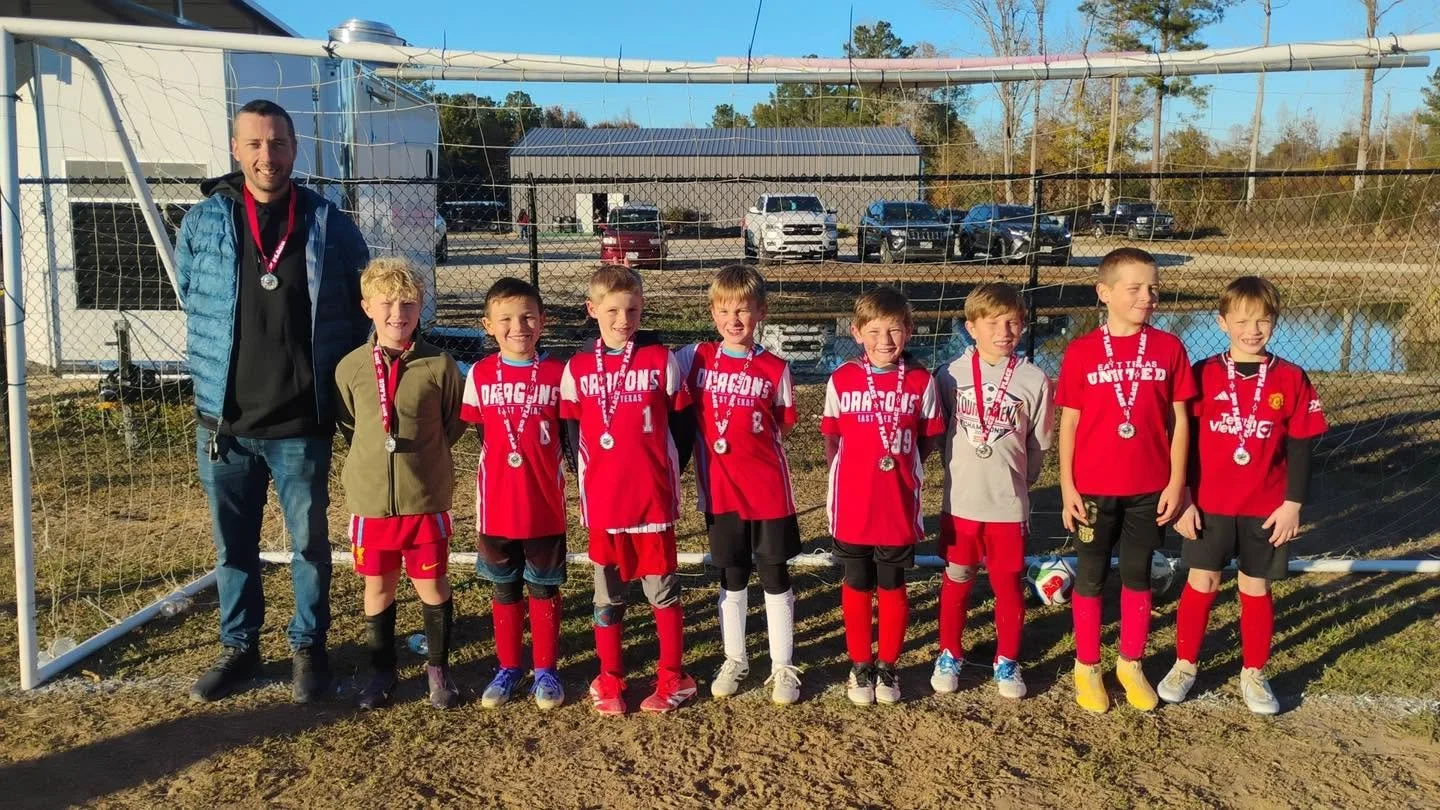 Group of young boys and their coach standing in front of a soccer goal on a field, all wearing medals, celebrating a game or tournament win, with soccer balls at their feet and parked cars in the background.
