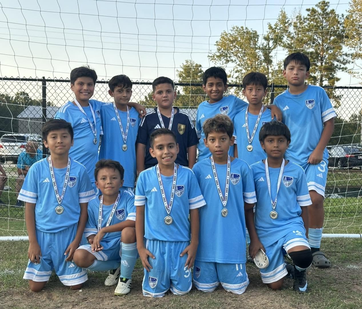 A group of young boys in light blue soccer uniforms with medals around their necks, posing in front of a soccer goal net on a grassy field during the daytime.