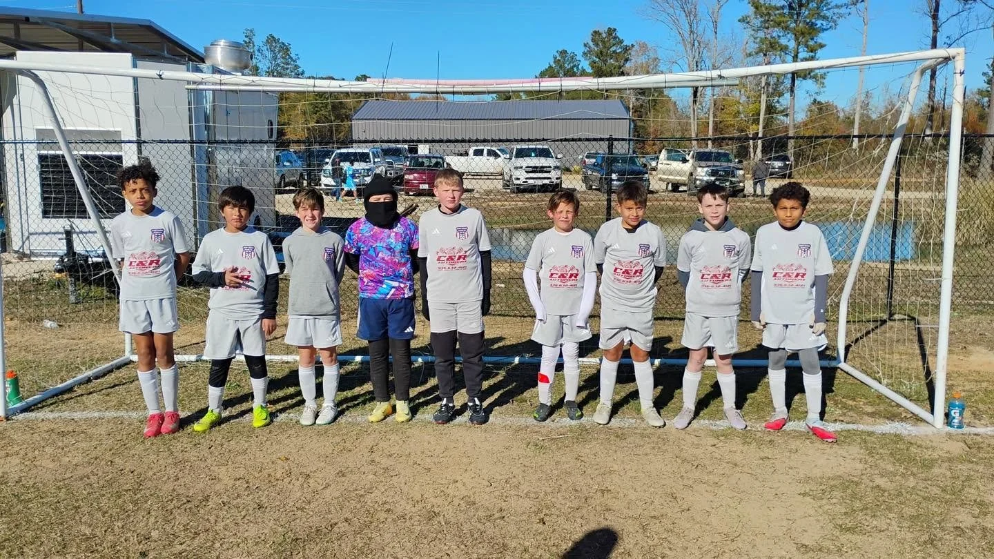 Young soccer team standing in front of a goal net during daytime. The team is wearing gray jerseys and shorts, with some players wearing black or white long sleeves underneath their jerseys. One player is wearing a colorful jacket and a black face mask. The background shows parked cars, trees, and a building with a blue sky above.