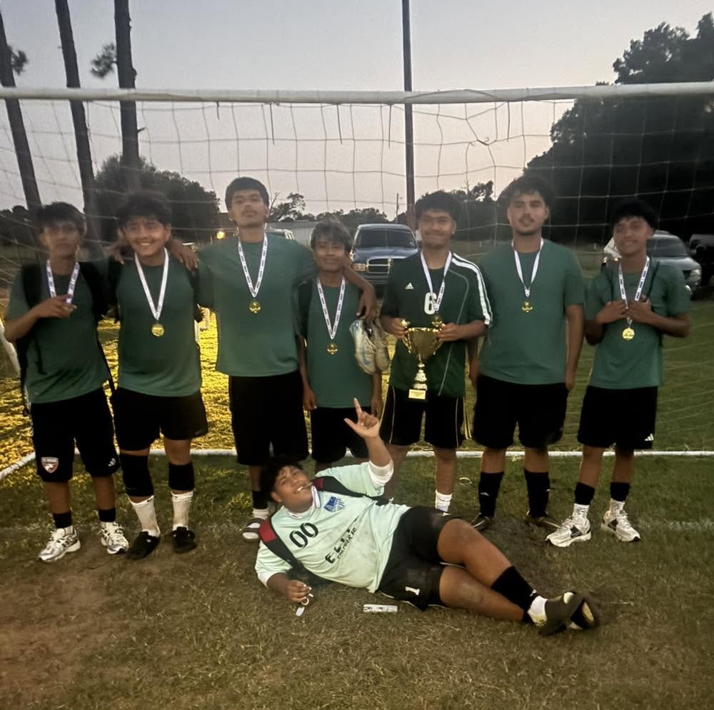 Group of young soccer players wearing medals, standing behind a soccer goalpost on a field, with a person lying on the ground in front, holding a trophy.