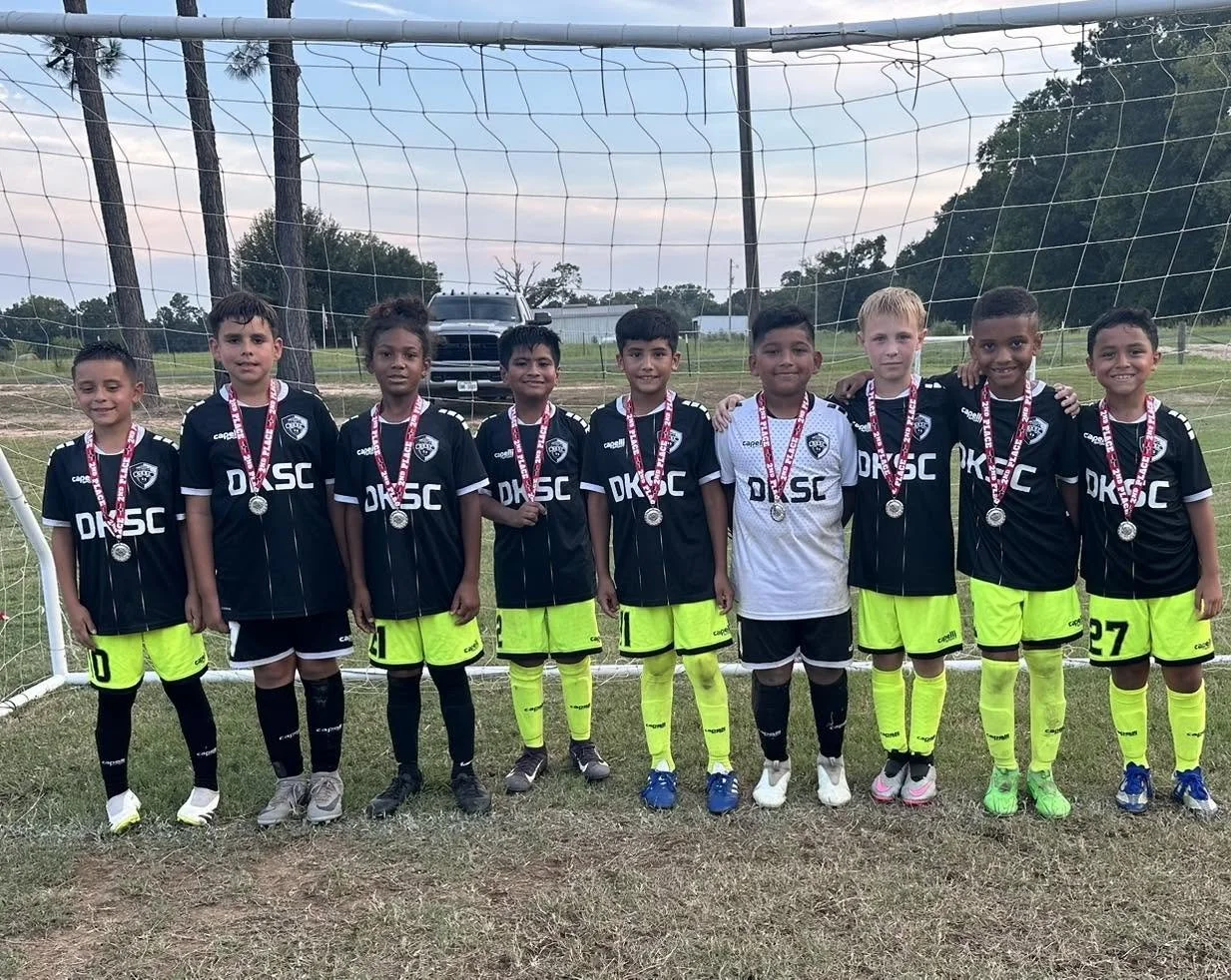 Young soccer players wearing black and neon yellow uniforms with medals, standing in front of a soccer goal on a grassy field.
