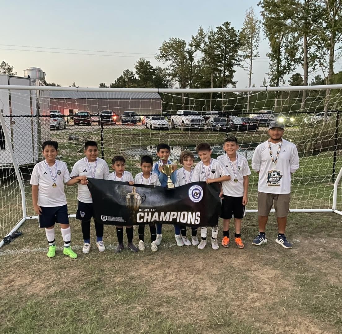 Group of young boys in soccer uniforms standing in front of a soccer goal, holding a banner that says 'We Are the Champions,' with some wearing medals and holding a trophy.