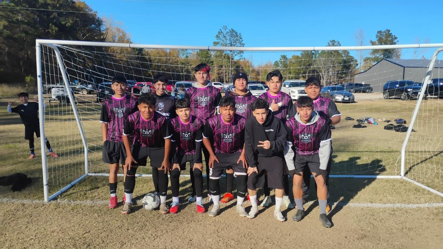 A group of ten young boys in black and pink soccer uniforms posing in front of a soccer goal on a field, with a boy in black standing to the side and parking lot in the background.