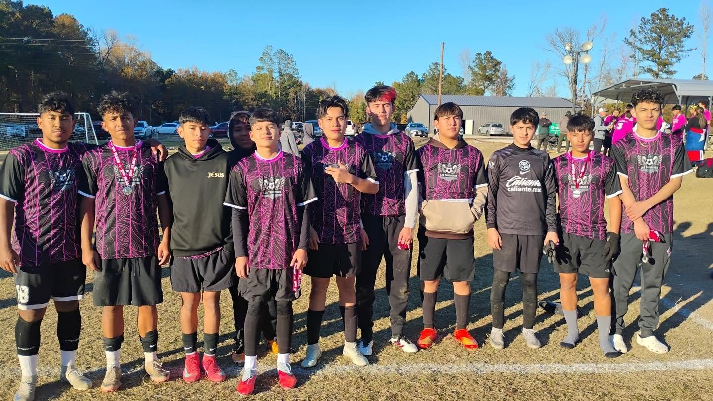 A group of young soccer players standing together on a soccer field after a game or practice, with some wearing medals, in sunny outdoor setting.