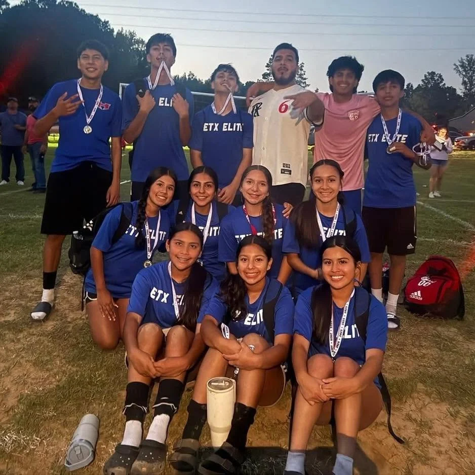 Group of young people, including boys and girls, celebrating on a soccer field. The girls are seated in the front and all are wearing blue team shirts, some with medals. The boys are standing behind, also with medals, some making poses. It is evening, and the background shows trees and other people.