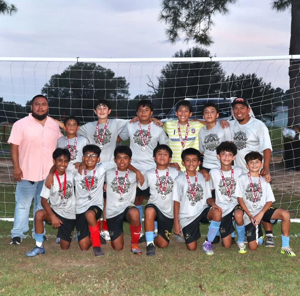 A youth soccer team of 11 boys with two adults, standing in front of a soccer goal on a field, wearing medals and posing for a group photo.
