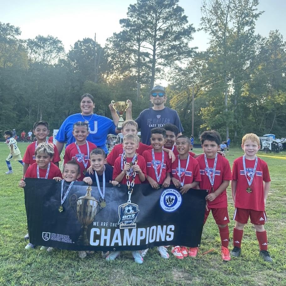 A group of young boys in red soccer uniforms holding medals, standing behind a banner that says 'We are the 903 Champions' with trophies, celebrating a victory on a soccer field, with adults standing behind them and sunlight in the background.
