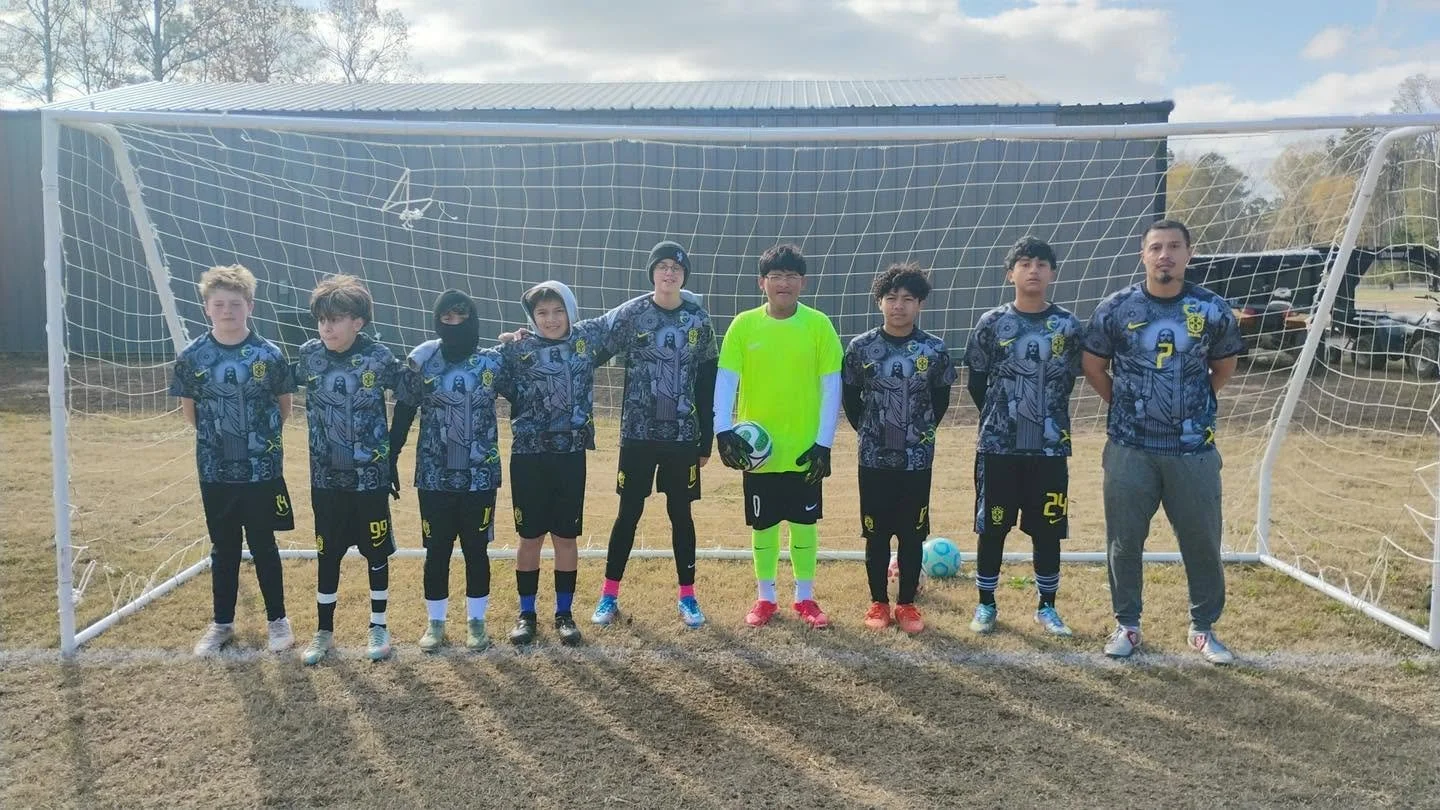 Group of young soccer players standing in front of a goal on a soccer field, wearing matching jerseys, with two adults, one goalkeeper holding a ball, and one dressed as a goalkeeper in a neon green kit, during daytime.